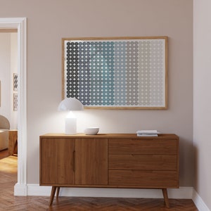 Op de afbeelding: Een houten dressoir in moderne mid-century stijl met twee deuren en drie lades. Het dressoir is lichtbruin van kleur en heeft een witte lamp en een kom erop. Het dressoir staat voor een lichtbeige muur met een ingelijst abstract print van blauwe en grijze cirkels.