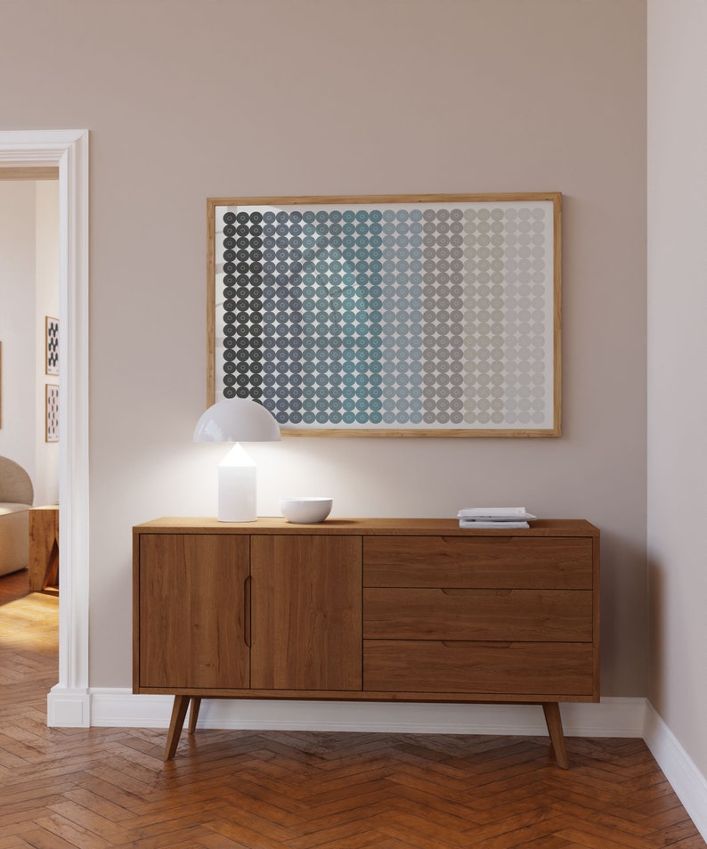 Op de afbeelding: Een houten dressoir in moderne mid-century stijl met twee deuren en drie lades. Het dressoir is lichtbruin van kleur en heeft een witte lamp en een kom erop. Het dressoir staat voor een lichtbeige muur met een ingelijst abstract print van blauwe en grijze cirkels.