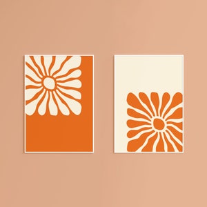 May include: Two framed prints with a white floral design on a bright orange background. The floral design is a stylized daisy with a large center circle.