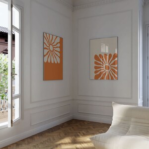 May include: Two framed abstract art prints with a white flower design on an orange background. The prints are hanging on a white wall in a room with a white couch.