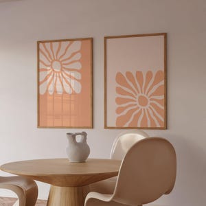 May include: Two framed prints with abstract floral designs in peach and white. The prints are hanging on a white wall above a wooden table with two white chairs.