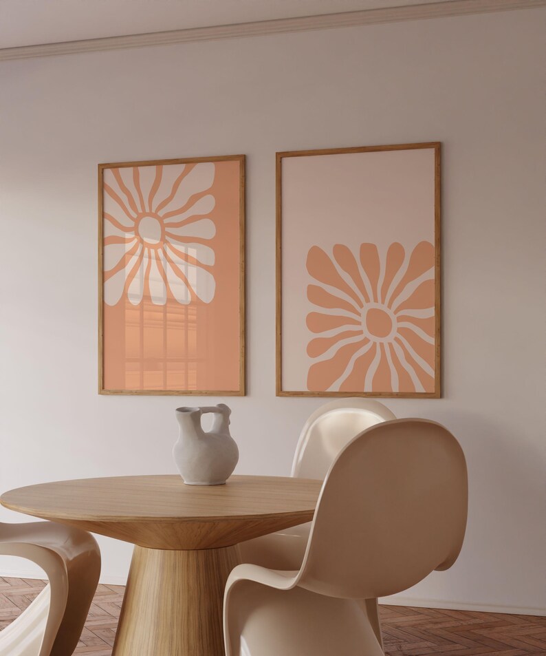 May include: Two framed prints with abstract floral designs in peach and white. The prints are hanging on a white wall above a wooden table with two white chairs.