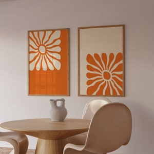 May include: Two framed prints with orange backgrounds and white stylized flower designs. The prints are hanging on a white wall above a wooden table with two white chairs.