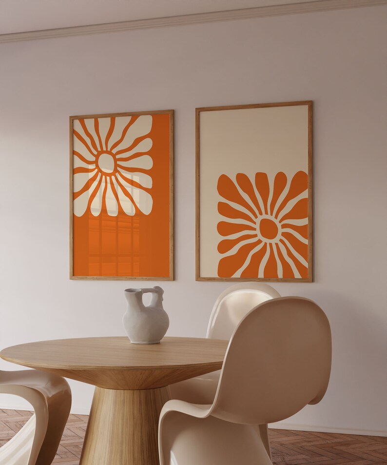 May include: Two framed prints with orange backgrounds and white stylized flower designs. The prints are hanging on a white wall above a wooden table with two white chairs.