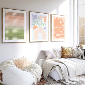 May include: Three framed abstract art prints in a living room setting. The prints feature a geometric pattern of circles in shades of pink, green, and brown, a floral design with a vase and rainbow, and a swirling abstract design in peach.