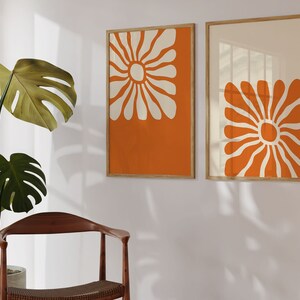 May include: Two framed prints with orange and white abstract floral designs. The prints are hung on a white wall.
