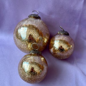 Puede incluir: Tres adornos navideños dorados con acabado craquelado. Los adornos son de diferentes tamaños, con una tapa de metal y un lazo para colgar. Los adornos reflejan la luz, creando un efecto brillante. El fondo es de color lavanda sólido.