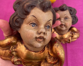 Może przedstawiać: Dwie antyczne rzeźby ścienne cherubinów. Cherubiny mają szczegółowe twarze z niebieskimi oczami i brązowymi kręconymi włosami. Cherubiny są pomalowane złotymi i czerwonymi akcentami. Tło jest w jasnoróżowym kolorze.