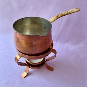 May include: A copper fondue pot with a woven handle and a stand. The pot has a rounded body and a silver-colored interior. The stand has a decorative design with curved legs and a circular base. A white candle is inside the stand.