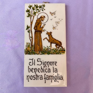 May include: A rectangular white ceramic plaque featuring a brown-robed figure with a halo standing near a tree and a wolf, with white birds flying above. The text "Il Signore benedica la nostra famiglia" is printed in black at the bottom.