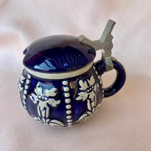 Może przedstawiać: Mały, kobaltowoniebieski ceramiczny kufel z metalową pokrywką na zawiasach i uchwytem. Kufel jest zdobiony białymi, kwiatowymi wzorami reliefowymi i białym paskiem wokół krawędzi. Pokrywka jest w ciemniejszym odcieniu niebieskiego.