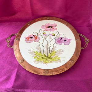 May include: A round copper serving tray with brass handles. The tray has a white ceramic insert with a painted design of pink and red poppies.