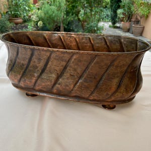 May include: An oval copper planter with a hammered finish and a decorative ribbed design. The planter has three feet and is made of a dark copper color.