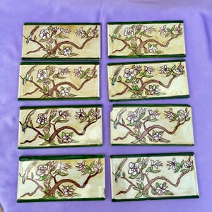 May include: Eight rectangular ceramic tiles with a hand-painted design. Each tile features a bird perched on a flowering branch with white blossoms and green leaves, set against a cream-coloured background with a green border.