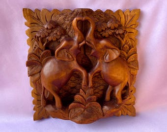 May include: A carved wooden wall plaque depicting two elephants with trunks touching to form a heart. The elephants are set within a detailed relief of leaves and branches. The artwork is framed with a decorative border.