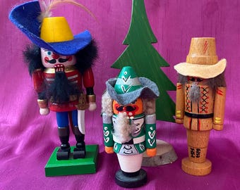May include: Three wooden nutcrackers with detailed painted features and hats. The nutcrackers are in various colors, including red, blue, green, and natural wood tones. A green wooden Christmas tree is in the background.