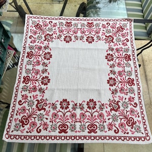 May include: White square tablecloth with a detailed floral design in shades of red, pink, and gray. The border features a repeating pattern of small loops. The floral pattern is concentrated around the edges.