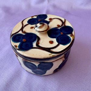 Puede incluir: Un tarro de cerámica redondo con tapa, con un diseño floral. El tarro es de color crema con patrones de flores azul oscuro y detalles marrones. La tapa tiene un pequeño pomo.
