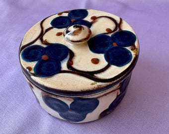 May include: A round ceramic jar with a lid, featuring a floral design. The jar is cream-colored with dark blue flower patterns and brown accents. The lid has a small knob and the jar is sitting on a purple surface.