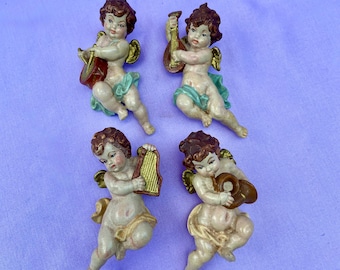 Puede incluir: Cuatro figuras de querubines vintage, cada una tocando un instrumento musical diferente. Los querubines tienen cabello castaño, alas doradas y visten ropa azul claro y beige. Los instrumentos incluyen un tambor, un laúd, un arpa y una trompa.