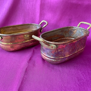 Può includere: Due ciotole ovali antiche in rame con manici in ottone. Le ciotole hanno una finitura anticata, che mostra sfumature di rame e bronzo. I manici sono curvi e attaccati ai lati. Le ciotole sono su uno sfondo viola.