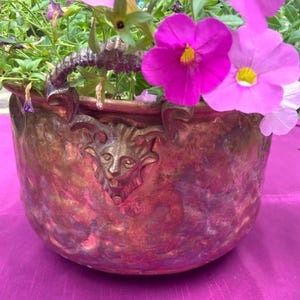 May include: A copper-toned planter with a detailed face design and handle, filled with vibrant pink and purple petunias. The planter has a weathered appearance, adding to its rustic charm. The flowers are in full bloom.