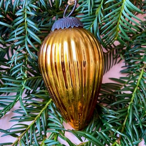 Op de afbeelding: Een goudkleurige, geribbelde glazen kerstversiering met een donkere metalen dop en een kleine lus om op te hangen. De versiering is druppelvormig en reflecteert licht. Het rust op een bedje van groene dennennaalden.