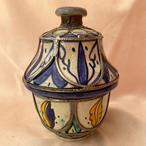 Op de afbeelding: Een keramische pot met een deksel, versierd met blauwe, witte, gele en groene bloemenpatronen. De pot heeft een zilveren metalen rand rond de boven- en onderkant.