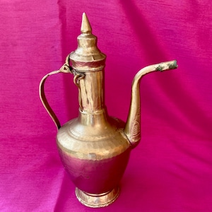 Puede incluir: Una jarra de latón vintage con un pico largo y curvado y un asa. La jarra tiene una textura martillada y una tapa cónica con un remate puntiagudo. El diseño sugiere una influencia antigua o de Oriente Medio. La jarra se muestra sobre un fondo rosa.
