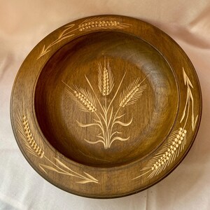 Op de afbeelding: Een ronde houten kom met een donkerbruine afwerking. De kom heeft gesneden tarwearen in het midden en langs de rand. Het tarwepatroon heeft een lichtere kleur dan het oppervlak van de kom. De kom wordt waarschijnlijk gebruikt om voedsel te serveren.