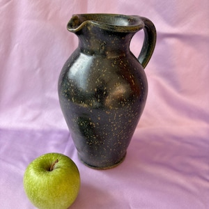 May include: A dark green ceramic pitcher with a handle and a ruffled rim. The pitcher has a speckled pattern and sits next to a green apple. The pitcher is approximately 9 inches tall.