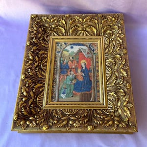 Op de afbeelding: Een goudkleurige, sierlijke fotolijst met een gedetailleerd bloemmotief. De lijst omringt een kleinere, rechthoekige afbeelding die een religieuze scène met figuren in kleurrijke gewaden weergeeft. De algehele uitstraling is vintage en decoratief.