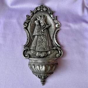 Può includere: Un'acquasantiera da parete color argento con un design ornato. L'immagine centrale raffigura una figura religiosa che tiene in braccio un bambino, incorniciata da un'elaborata cornice decorativa. L'acquasantiera ha un bacino in basso.