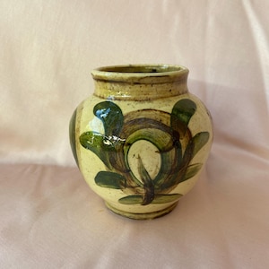 Può includere: Un piccolo vaso rotondo in ceramica con una glassa giallo chiaro e disegni floreali dipinti in verde e marrone.