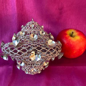 Op de afbeelding: Een zilverkleurig, versierd, decoratief object met een traliewerkontwerp en meerdere heldere strass steentjes. Het object ligt op een roze oppervlak naast een rode appel.