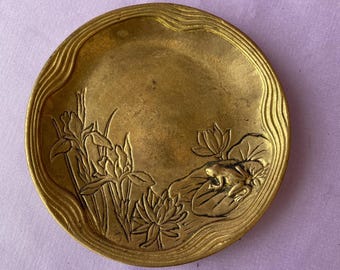 May include: A round, antique brass dish with a detailed relief design. The design features aquatic plants, including irises and water lilies, and a small frog on a lily pad. The dish has a wavy border.