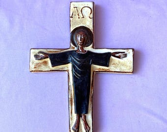 May include: A ceramic cross with a figure in a dark robe, arms outstretched. The cross is off-white with brown accents, and the figure has a dark halo. The top of the cross has the Greek letters Alpha and Omega.