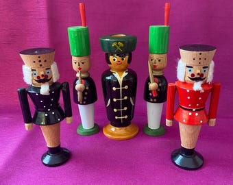 May include: Five wooden nutcrackers in various uniforms. The nutcrackers have painted details, including white beards and mustaches, and are dressed in black, red, and green outfits. Each nutcracker has a unique hat and holds a small object.