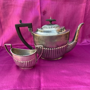 May include: A silver teapot and creamer set with a black handle on the teapot. The teapot has a fluted design and a silver knob on the lid. The creamer is also fluted and has two handles.