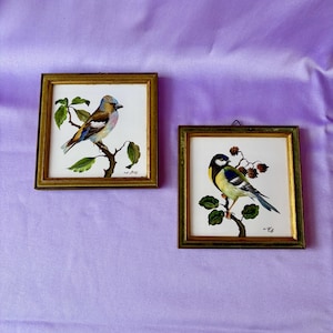 Puede incluir: Dos pinturas de pájaros enmarcadas. Una presenta un pájaro azul, marrón y amarillo posado en una rama con hojas verdes. La otra muestra un pájaro amarillo, negro y azul en una rama con bayas y hojas verdes. Ambas pinturas tienen marcos dorados.