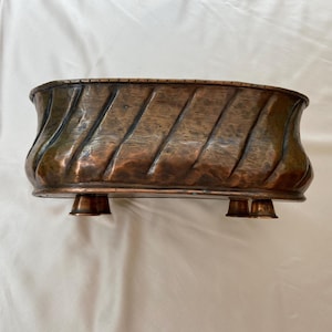 Puede incluir: Un macetero ovalado de cobre con un acabado martillado y un patrón de espiral decorativo. El macetero tiene tres patas y es perfecto para exhibir plantas.