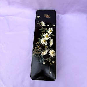 Könnte beinhalten: Eine rechteckige schwarze Lackschachtel mit einem bemalten Blumenmuster aus weißen Gänseblümchen mit grünen Stielen und Blättern. Ein kleiner brauner Schmetterling ist oben auf dem Deckel abgebildet. Die Oberfläche hat ein glattes, glänzendes Finish.