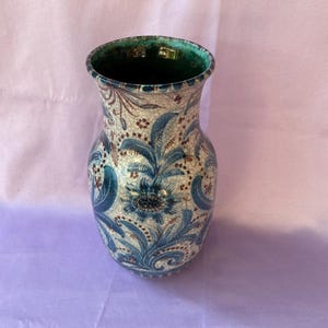 Puede incluir: Un jarrón de cerámica con acabado craquelado, con diseños florales azules sobre fondo crema. El jarrón tiene interior verde y borde acampanado. El diseño incluye flores y hojas estilizadas.