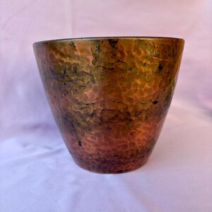 May include: A tapered, cylindrical vessel crafted from hammered copper, featuring a rich, multi-tonal patina that transitions from deep reddish-brown at the base to dark, oxidised green and bronze hues toward the upper rim.