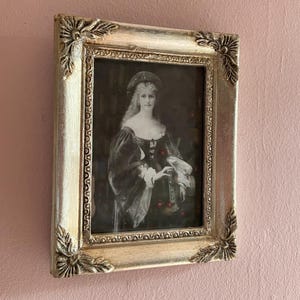 Puede incluir: Un retrato en blanco y negro enmarcado en plata de una mujer con un vestido y sombrero oscuros. El marco tiene esquinas decorativas. El retrato está montado en una pared rosa.