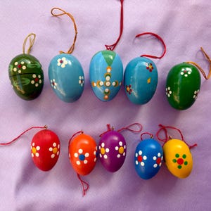 Puede incluir: Una colección de diez adornos de huevos de Pascua de madera pintados a mano en varios colores. Cada huevo presenta un diseño floral único con detalles blancos y amarillos. Los huevos están suspendidos por cuerdas rojas y doradas.