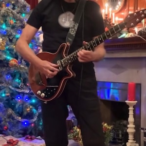 Può includere: Una persona suona una chitarra elettrica marrone e nera. La chitarra ha una forma e un design unici. La persona indossa una maglietta e pantaloni neri. Il testo sull'immagine dice: "This is a tease... For something nice which is coming in January."