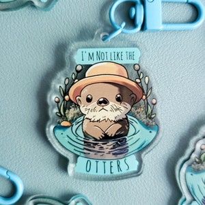 Könnte beinhalten: Ein blauer und weißer Acryl-Schlüsselanhänger mit einem Cartoon-Otter, der einen gelben Hut trägt und den Text "Ich bin nicht wie die Otter".