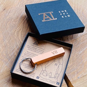 May include: A copper metal keychain, engraved with the Roman numeral VII, presented in a black box. The box features the text "SHEFFIELD" and the initials "AL" in copper. The keychain is displayed on a wooden surface.