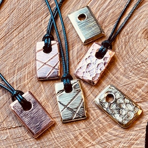 Op de afbeelding: Rechthoekige metalen hangerkettingen in verschillende afwerkingen, waaronder koper en goud. Elke hanger is bevestigd aan een zwart koord. De hangers hebben texturen of gegraveerde ontwerpen.
